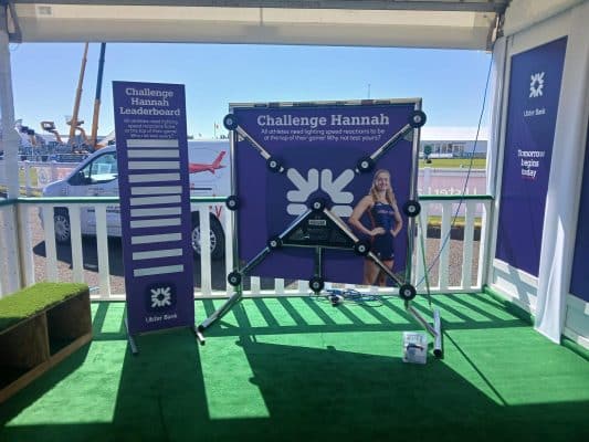 Purple 'Challenge Hannah' leaderboard display at an event with a metal arm-frame and a smiling woman in athletic wear beside it.
